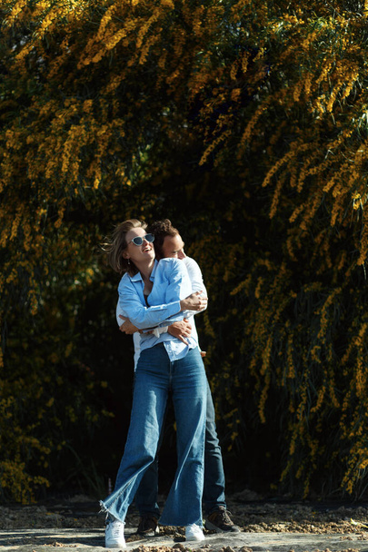 Foto al aire libre de pareja joven enamorada caminando por el camino a través del campo. Hombre y mujer caminando a lo largo de árboles de flores amarillas.  - Foto, imagen