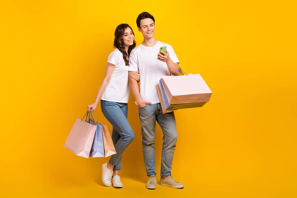 Full length photo of pretty positive couple wear white t-shirts holding bargains typing device space isolated yellow color background. - Foto, Bild