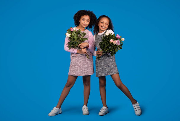 Cheerful cute black little girls girlfriends with bouquets of flowers, enjoy greeting birthday, isolated on blue background, studio. Holiday celebration, congratulation, childhood and emotions - Photo, Image