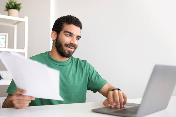 Smiling young middle eastern guy blogger at workplace with laptop works with documents in room office interior, free space. Freelance, accounting, success business and study remotely at home - Foto, Bild