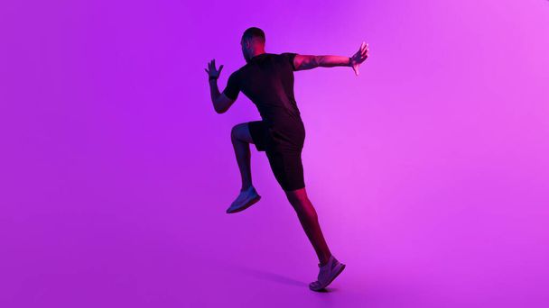 Trainingskonzept. Rückansicht des Afroamerikaners, der läuft und trainiert, schwarze Sportbekleidung auf lila Hintergrund trägt. Nicht wiederzuerkennen Fitness Guy Jumping Training im Studio. Panorama - Foto, Bild
