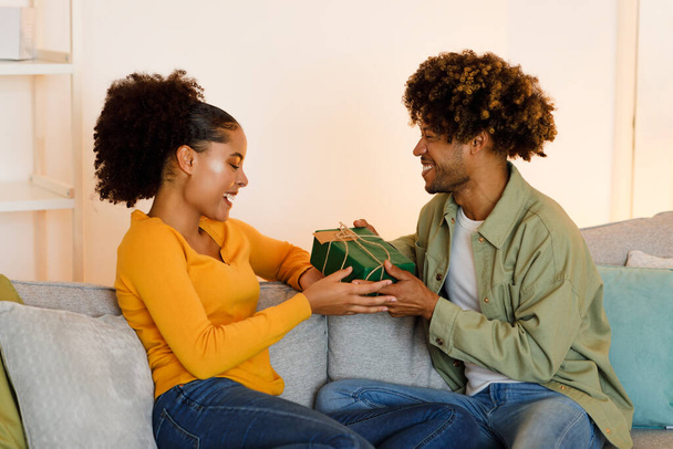 ロマンチックな贈り物だ。黒人夫へのプレゼント自宅でカウチに座っておめでとう妻を与えます.家族の休日や関係の記念日を祝う若いカップル一緒に - 写真・画像