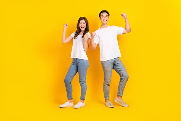 Full length photo of excited lucky couple wear white t-shirts rising fists empty space isolated yellow color background. - Foto, afbeelding