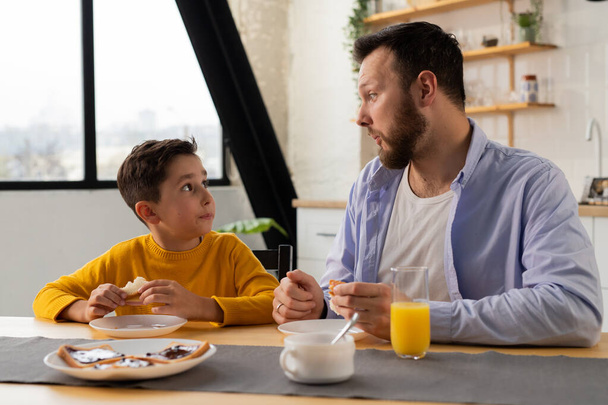 Vader en zoon zitten aan de keukentafel en kijken elkaar aan. De jongen zit aan tafel met zijn vader en eet sandwiches. Hoge kwaliteit foto - Foto, afbeelding