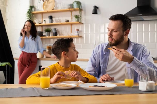 De jongen communiceert met zijn vader tijdens het ontbijt in de keuken. Vader en zoon kijken elkaar aan tijdens het ontbijt in de keuken. Hoge kwaliteit foto - Foto, afbeelding