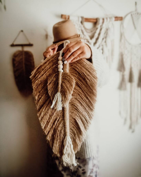 A vertical shot of a woman holding a macrame feather and a hat with a white wall in the background - Fotografie, Obrázek