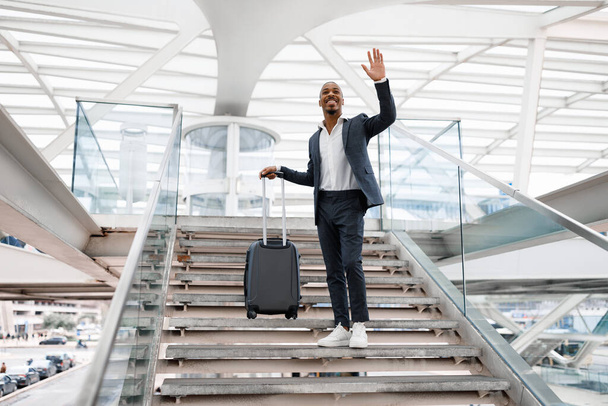 Glimlachende zwarte zakenman staande op trappen op de luchthaven Terminal en zwaaiende hand, Knappe Afro-Amerikaanse Man begroet iemand na aankomst, het dragen van koffer en op zoek naar weg, kopieerruimte - Foto, afbeelding