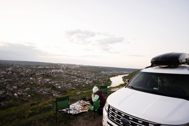 Woman  sit on chair, hiking on vacation, having evening picnic on top of the mountain, looking at beautiful canyon landscape. Car with roof rack box. - 写真・画像
