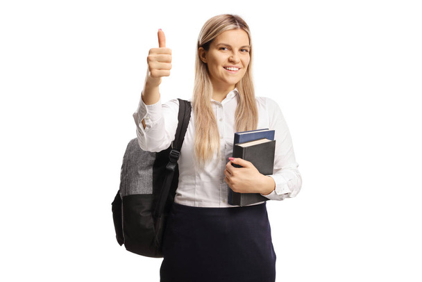 Cheeful female student holding books and showing a thumb up sign isolated on white background - Foto, Imagem