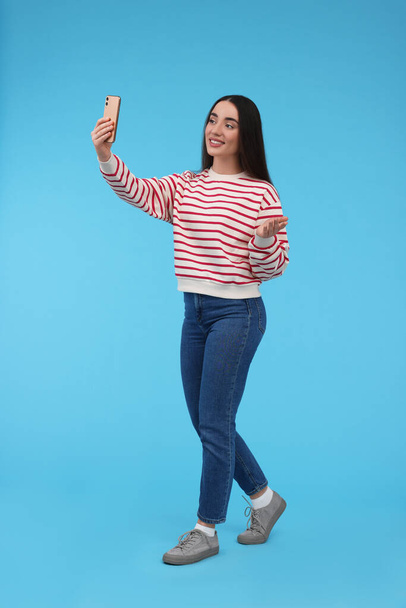 Smiling young woman taking selfie with smartphone on light blue background - Zdjęcie, obraz