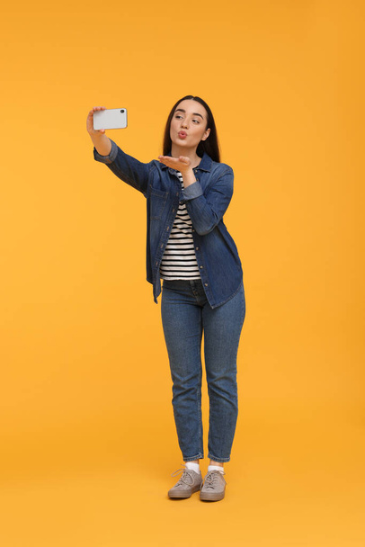 Young woman taking selfie with smartphone and blowing kiss on yellow background - Fotó, kép