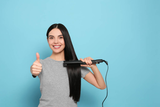 Beautiful happy woman showing thumbs up while using hair iron on light blue background. Space for text - Valokuva, kuva