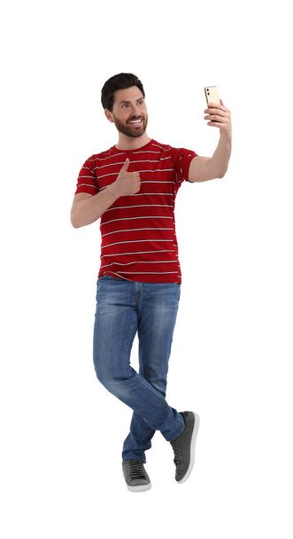 Smiling man taking selfie with smartphone and showing thumbs up on white background - Photo, Image