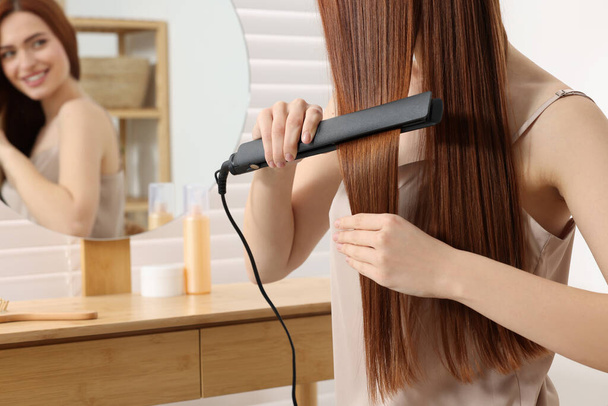 Beautiful woman using hair iron near mirror in room, closeup - Φωτογραφία, εικόνα