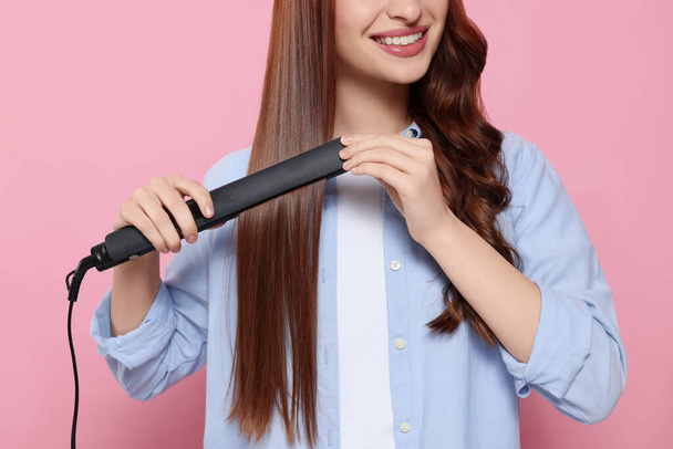 Young woman using hair iron on pink background, closeup - Foto, afbeelding
