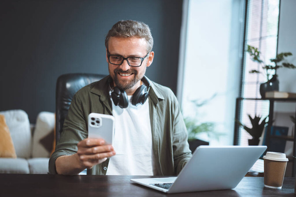 Happy and smiling man is sitting in a loft office, dressed in casual wear, working on his laptop while texting a message on his phone. High quality photo - Foto, Imagem