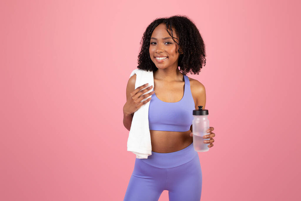 Gülümseyen Afro-Amerikan kıvırcık kadın havluyla ve bir şişe suyla pembe stüdyo arka planında eğleniyor. Formda yaşam tarzı, vücut bakımı, aktif ve egzersiz - Fotoğraf, Görsel
