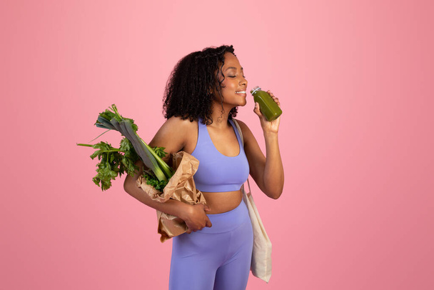 Cheerful young african american curly lady in sportswear hold bag of vegetables and drinking bottle of smoothie, isolated on pink studio background. Fit lifestyle, body care, diet and healthy food - Foto, afbeelding