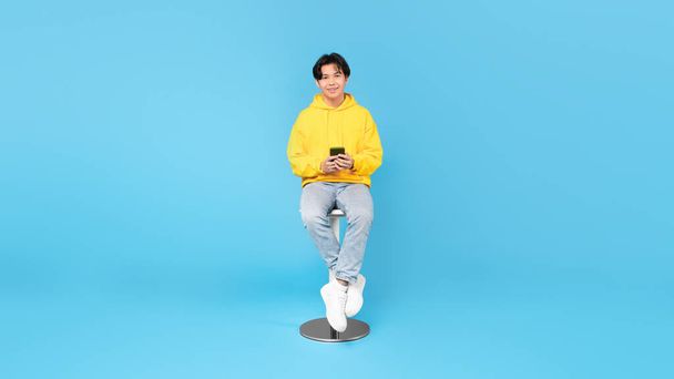 Új mobil alkalmazás. Boldog kínai tinédzser srác használ okostelefon sms-ezés és böngészés Internet ül a szék felett kék háttér. Szerkentyűk Ajánlat koncepció. Panoráma, Stúdió lövés - Fotó, kép