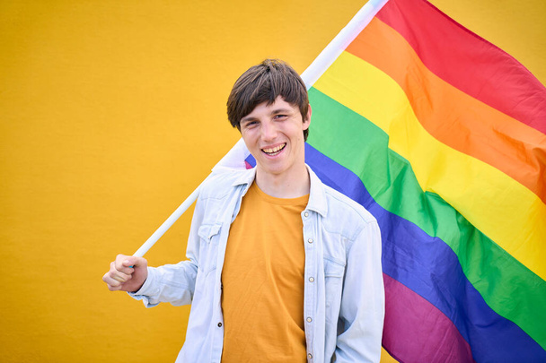 Uśmiechnięty młody biały trzymający tęczową flagę Igbt. Homoseksualiści pozujący do zdjęć z żółtym tłem. Wesoły gej człowiek szczęśliwy studio zdjęcie na izolatce. Pokolenie z. - Zdjęcie, obraz