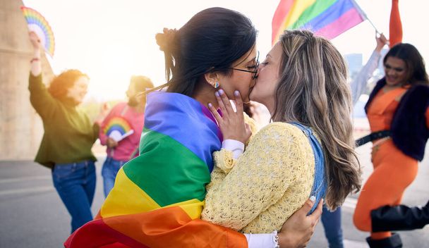 Dos jóvenes chicas cariñosas besándose en el día del festival del orgullo gay al aire libre. Grupo de amigos celebrando fiesta lgbt en el fondo con la bandera del arco iris y los fans. Generación z y tipos de sexualidad. - Foto, Imagen