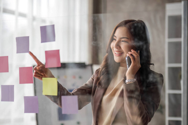 Young smiley attractive, businesswoman using sticky notes in glass wall to writing strategy business plan to development grow to success.. - 写真・画像