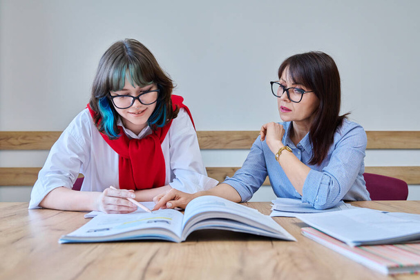 Young teenage female studying foreign languages in course with teacher. Tutor and student sitting at desk in office, using books, notebooks. Education, knowledge, learning concept - Photo, Image