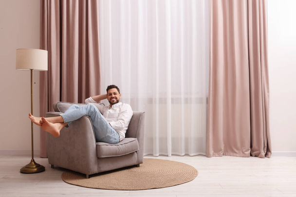 Happy man resting on armchair near window with beautiful curtains at home. Space for text - Fotoğraf, Görsel