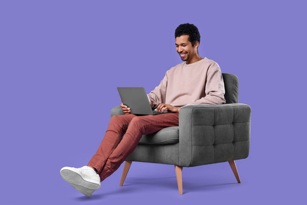 Happy man with laptop sitting in armchair on purple background - Foto, immagini