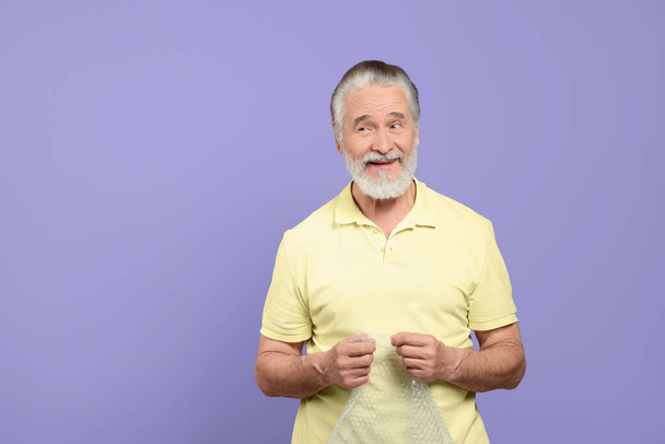 Happy senior man popping bubble wrap on light purple background, space for text. Stress relief - Photo, Image