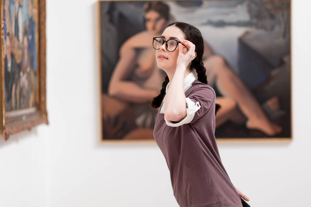 Cultural education. Side view of caucasian young woman student wearing glasses visiting excursion in museum, contemplates arts. Concept of exhibition in gallery. - Fotó, kép