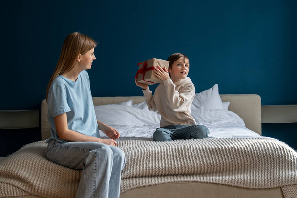 Happy curious little boy sitting on bed holding wrapped gift box guessing what is inside, excited kid getting birthday present from loving mom at home. Mother congratulating son, making surprise - Photo, Image