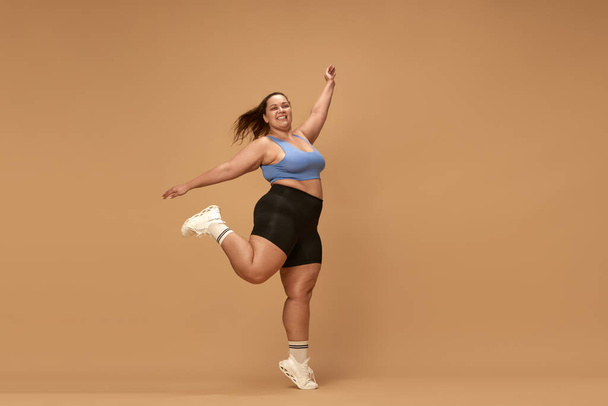 Young, active, smiling, overweight woman training in sportswear against brown studio background. Workout with positivity. Concept of sport, body-positivity, weight loss, body and health care - Photo, image