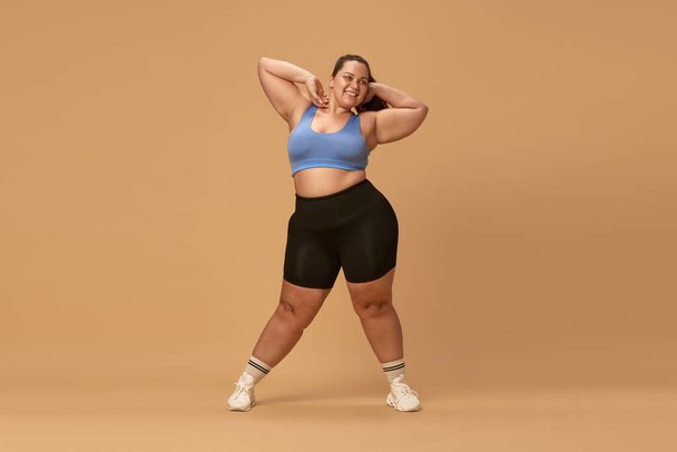 Smiling, positive, young overweight woman posing in comfortable sportswear against brown studio background. Caring about body. Concept of sport, body-positivity, weight loss, body and health care - Foto, imagen