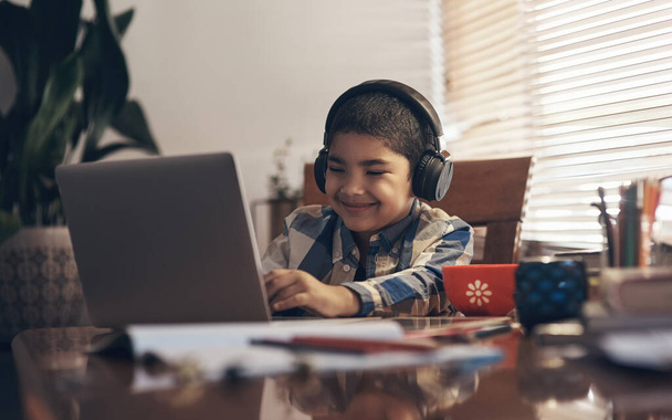 Najfajniejsza szkoła na świecie. uroczy chłopiec korzystający z laptopa i słuchawek podczas wykonywania zadania szkolnego w domu - Zdjęcie, obraz