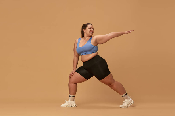 Young smiling woman with fat, overweight body training in sportswear against brown studio background. Active lifestyle. Concept of sport, body-positivity, weight loss, body and health care - Фото, изображение