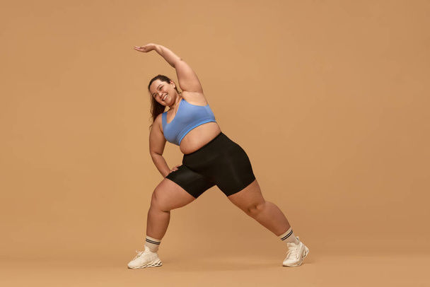 Stretching. Young overweight woman training in sportswear against brown studio background. Motivation to workout. Concept of sport, body-positivity, weight loss, body and health care - Photo, Image