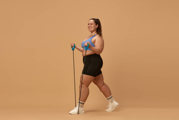 Young overweight woman smiling, training in sportswear with fitness equipment, expanders against brown studio background. Concept of sport, body-positivity, weight loss, body and health care - Photo, Image