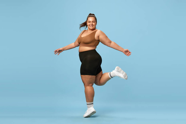 Young, happy, active, overweight woman training in sportswear against blue studio background. Self-love and acceptance. Concept of sport, body-positivity, weight loss, body and health care - Foto, Bild