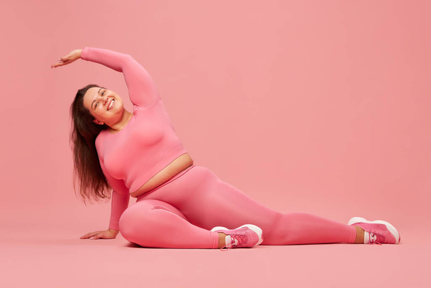 Well-being and self-love. Young overweight woman training in pink sportswear, doing stretching against pink studio background. Concept of sport, body-positivity, weight loss, body and health care - Foto, Imagem