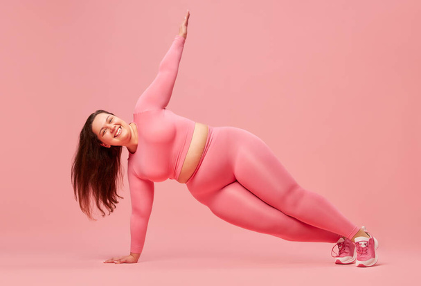 Young, positive, motivated woman with overweight body training in sportswear against pink studio background. Exercising. Concept of sport, body-positivity, weight loss, body and health care - Foto, Bild