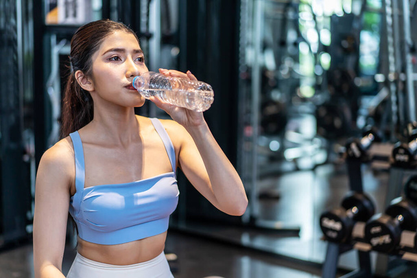 Portret zmęczonej Azjatki w odzieży sportowej pijącej wodę z butelki na siłowni. Samica sportowca uzupełniająca równowagę wodną po treningu fitness. pragnienie po treningu lub treningu fizycznym - Zdjęcie, obraz