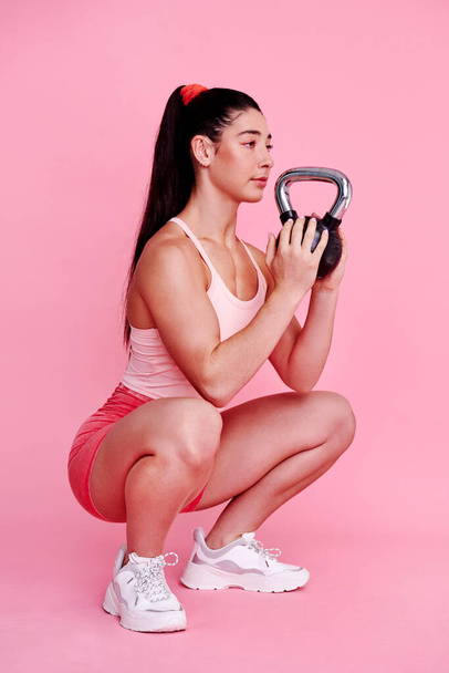 Those who work hard earn the best rewards. Studio shot of a sporty young woman doing squats with a kettlebell against a pink background - Fotoğraf, Görsel