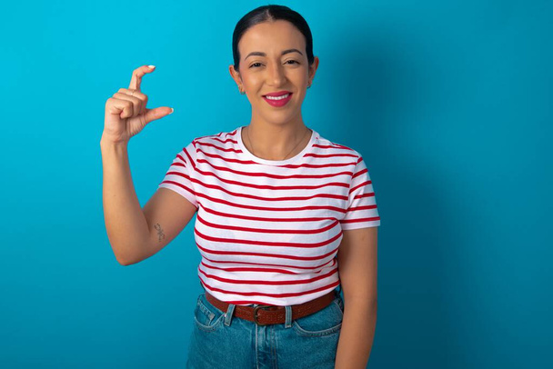 mujer vistiendo camiseta a rayas sonriendo y haciendo gestos con la mano de tamaño pequeño, símbolo de medida - Foto, imagen
