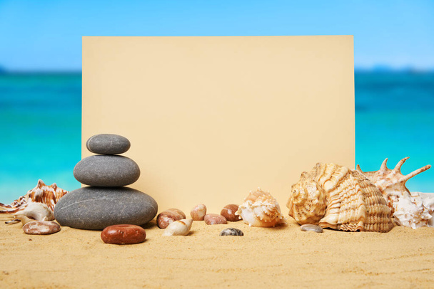 Invitation or greeting card mockup with seashells and pyramid of stones on the summer sandy beach at ocean background. Vacation concept, copy space - Fotó, kép