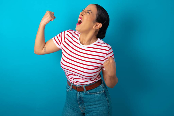 Attractive woman wearing striped T-shirt celebrating a victory punching the air with his fists and a beaming toothy smile - Photo, image