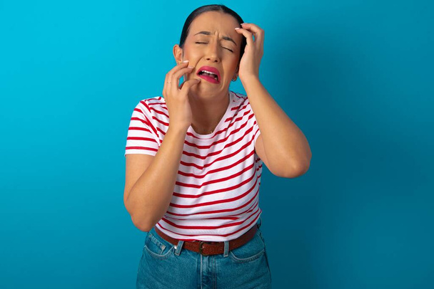 Young gloomy woman wearing striped T-shirt , hiding face with hands pouting and crying, standing upset and depressed complaining about job problem. - Photo, Image