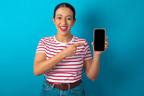 Smiling woman wearing striped T-shirt showing and pointing at empty phone screen. Advertisement and communication concept. - Фото, изображение