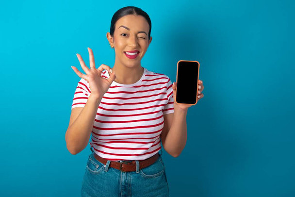 Excited woman wearing striped T-shirt showing smartphone blank screen, blinking eye and doing ok sign with hand.  Advertisement concept. - Φωτογραφία, εικόνα