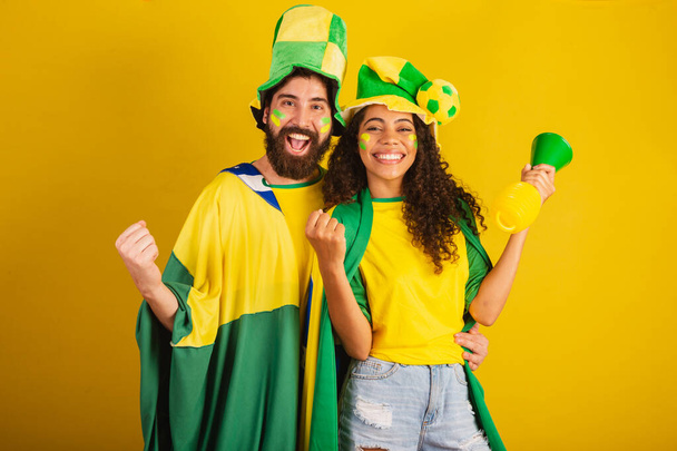 couple of brazil soccer supporters, dressed in the colors of the nation, black woman, caucasian man. using flag and accessories - Foto, Imagen